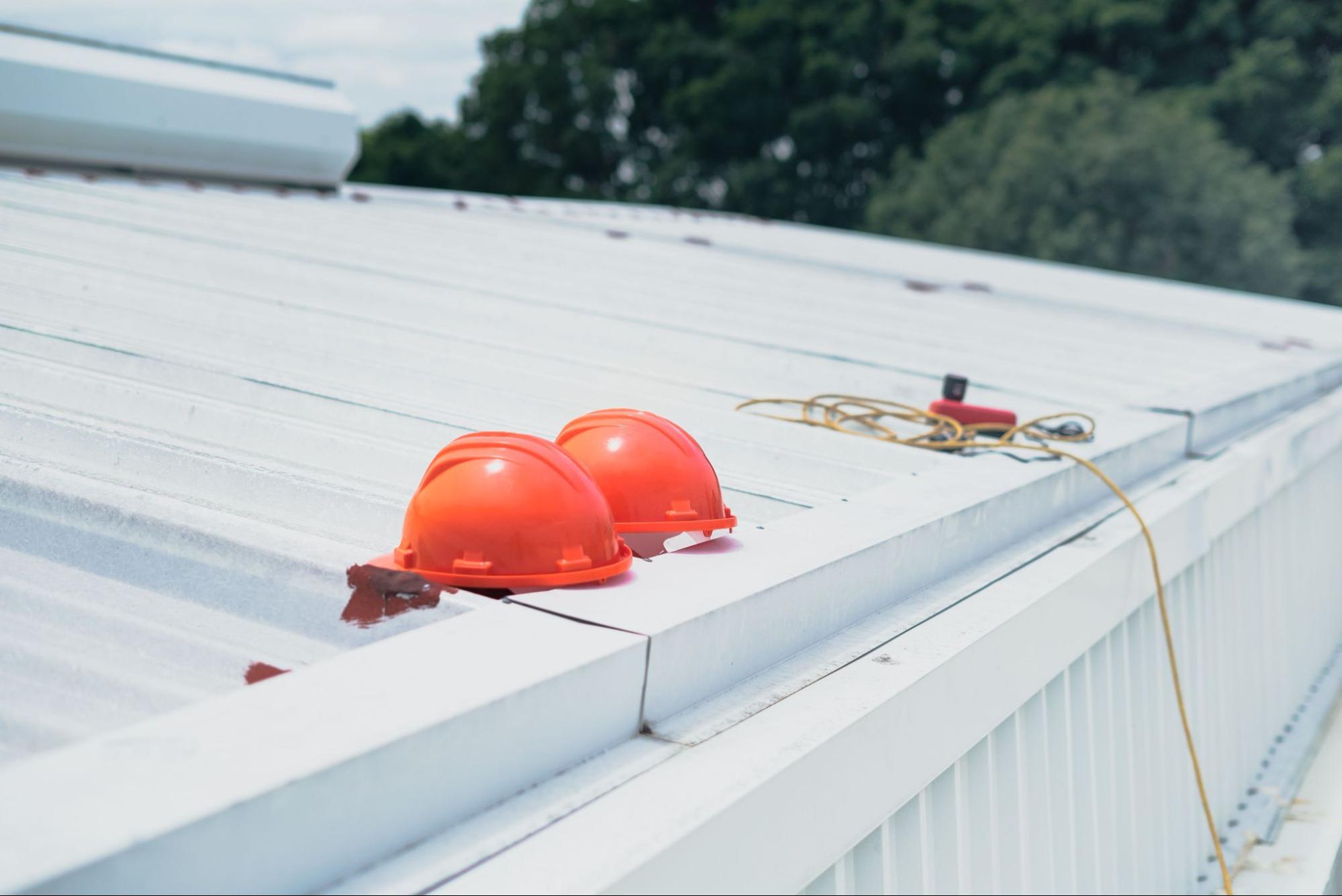 Hard hats resting on a metal commercial roofing panel beside an edge guardrail.